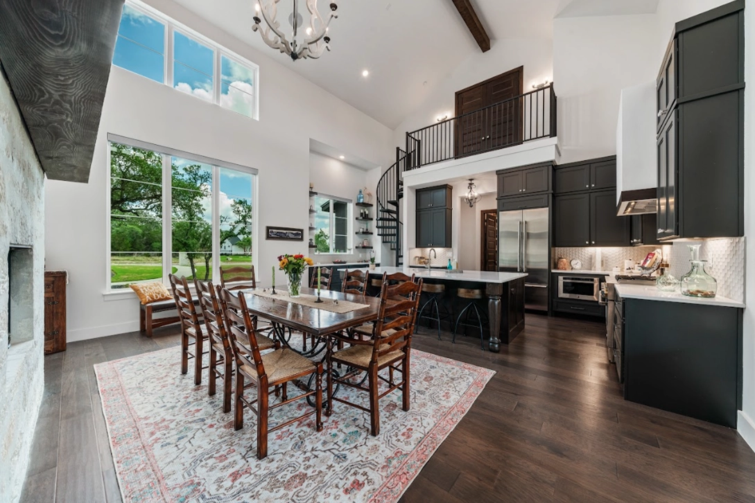 Spacious open-concept area combining a dining room and kitchen, featuring dark cabinetry, a large dining table, high ceilings, and large windows with a scenic view.